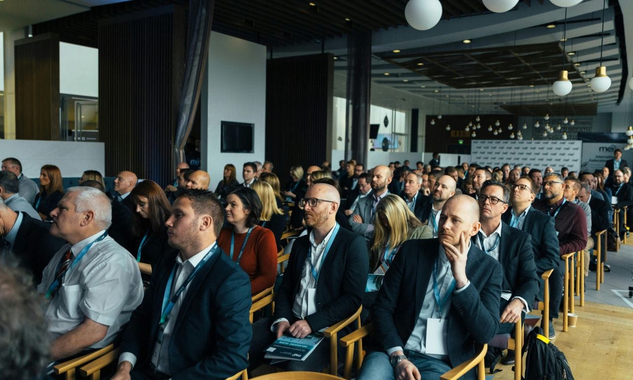 The image shows a group of people seated in an event or conference hall, all paying attention to a presentation or speaker. The attendees, dressed in formal or business attire, appear engaged, but their expressions indicate that their specific needs or interests might not be fully addressed in the session. This ties into the topic of "Ignoring Audience Needs" in event planning, where understanding and catering to the audience's expectations is crucial for a successful event.