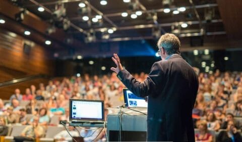 A speaker in a dark suit delivers a presentation to a large audience in a modern conference hall. A laptop displaying slides is placed on a podium, and the audience is attentively listening. The event appears to be a budget-friendly corporate gathering with a professional yet simple setup.