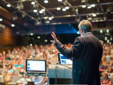 A speaker in a dark suit delivers a presentation to a large audience in a modern conference hall. A laptop displaying slides is placed on a podium, and the audience is attentively listening. The event appears to be a budget-friendly corporate gathering with a professional yet simple setup.